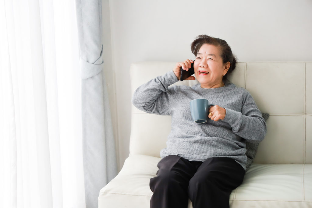 Asian senior woman talking on a mobile phone and drinking coffee in living room.
